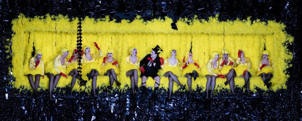 Row of women with a man in the middle against a yellow backdrop - Photo from The Cunning Little Vixen opera.