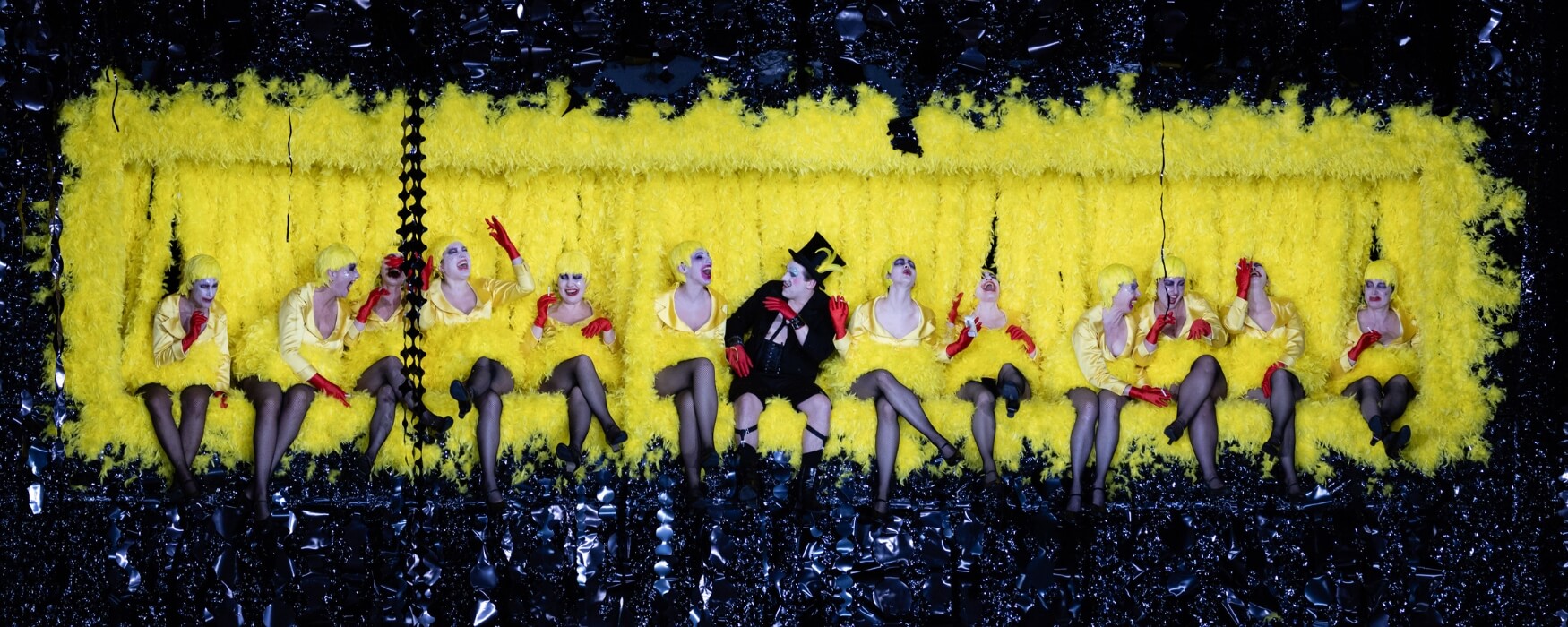 Row of women with a man in the middle against a yellow backdrop - Photo from The Cunning Little Vixen opera.