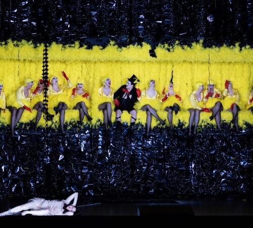 Row of women with a man in the middle against a yellow backdrop - Photo from The Cunning Little Vixen opera.