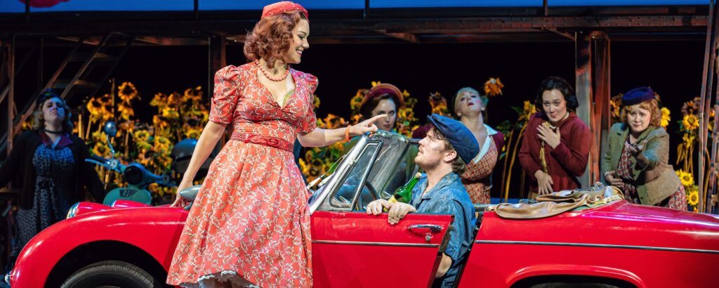 Woman in red dress leaning against a vintage red car facing the man driving. Photo from The Elixir of Love opera.