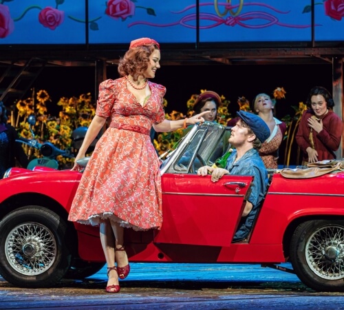 Woman in red dress leaning against a vintage red car facing the man driving. Photo from The Elixir of Love opera.