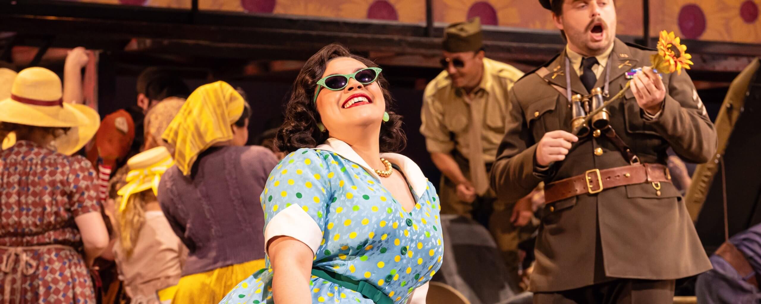 Woman in a blue dress and sunglasses singing with a man in a uniform carrying a sunflower - The Elixir of Love at The Dallas Opera